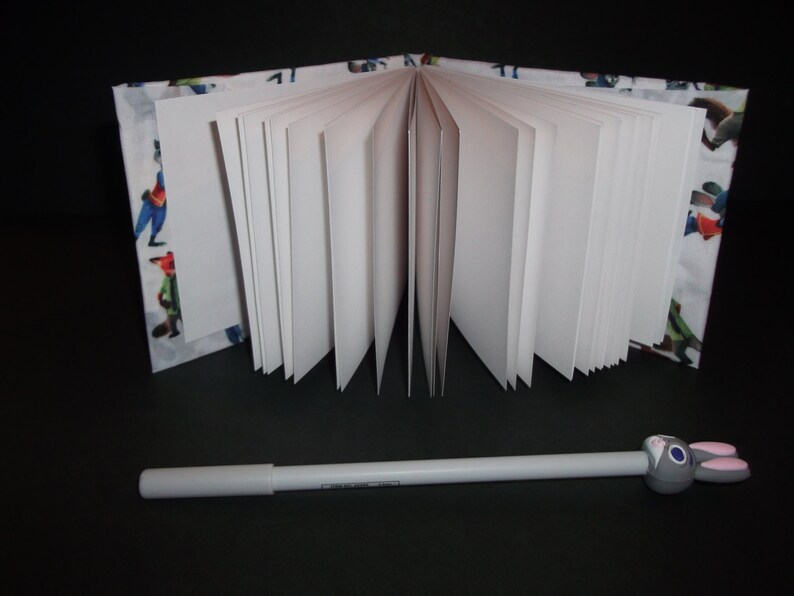 Peut inclure: Un cahier ouvert avec des pages blanches et une couverture &agrave; motifs avec des personnages de dessins anim&eacute;s color&eacute;s. Un stylo blanc avec un dessus en forme de lapin gris repose devant le cahier sur une surface noire.