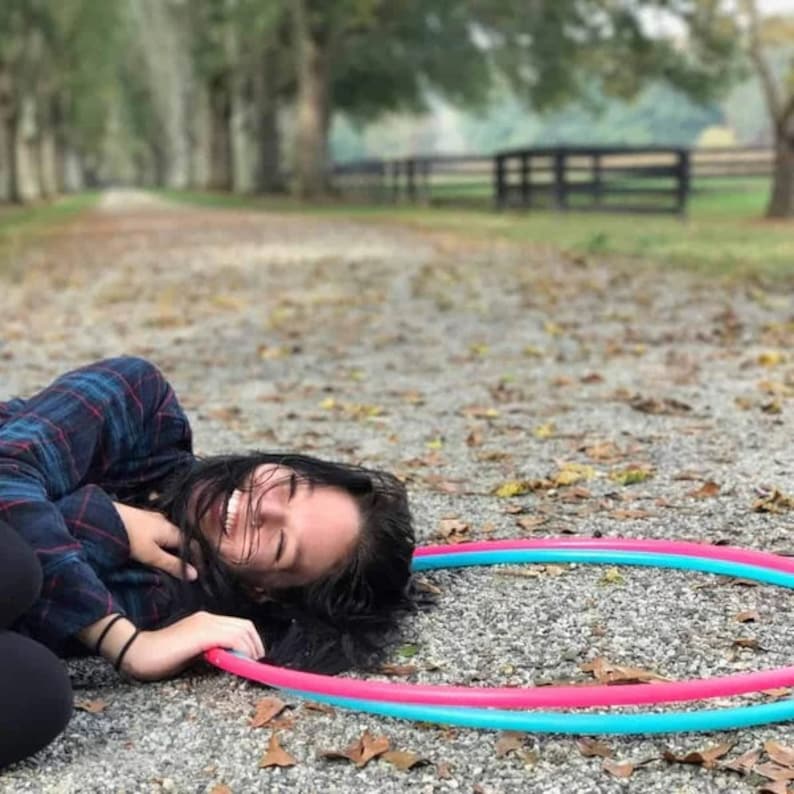 Op de afbeelding: Een vrouw ligt op haar rug op een grindpad en lacht. Ze draagt een geruit overhemd en heeft lang donker haar. Twee kleurrijke plastic hoepels liggen naast haar op de grond.