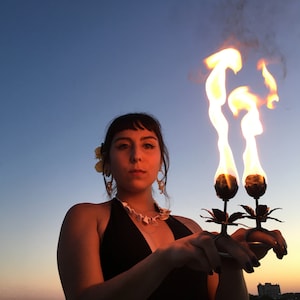 May include: A person holding two metal torch holders with flames coming out of the top. The torches are shaped like flowers. The person is wearing a black halter top and a flower behind their ear.