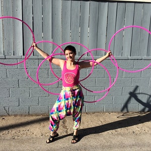May include: A person is holding five pink hula hoops in front of a gray brick wall. The person is wearing a pink tank top with a geometric design and colorful patterned pants.