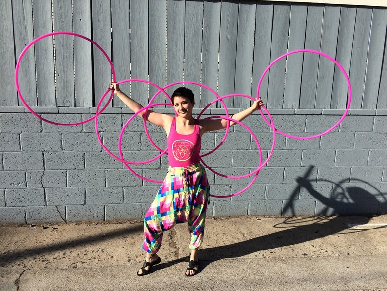 May include: A person is holding five pink hula hoops in front of a gray brick wall. The person is wearing a pink tank top with a geometric design and colorful patterned pants.