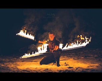 May include: A person in a dark outfit is performing a fire dance with two flaming staffs in a snowy setting. The person is holding the staffs in a crossed position, creating a fiery X shape.