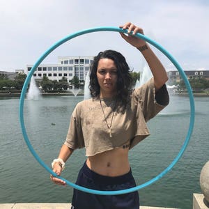 May include: A person is holding a blue plastic hula hoop in front of a body of water with fountains. The person is wearing a brown crop top and blue jeans.