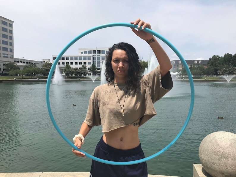 May include: A person is holding a blue plastic hula hoop in front of a body of water with fountains. The person is wearing a brown crop top and blue jeans.