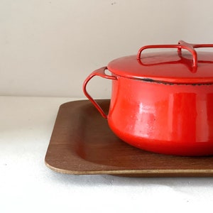 Puede incluir: Una olla de esmalte rojo brillante con tapa y asa a juego, colocada sobre una bandeja rectangular de madera. La olla tiene un diseño clásico, ideal para cocinar. La bandeja es de color marrón cálido.