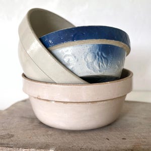 May include: A stack of three ceramic mixing bowls. The bottom bowl is a solid light beige. The middle bowl features a deep blue upper band with a textured floral pattern. The top bowl is a plain, light beige ceramic with a smooth, glossy finish.