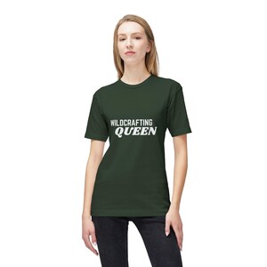May include: A dark forest green short-sleeved t-shirt featuring the white text "WILDCRAFTING QUEEN" printed across the chest. The shirt has a crew neckline and a standard fit, paired with dark grey jeans, creating a simple and casual appearance.