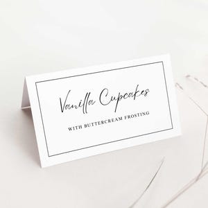 Könnte beinhalten: Eine gefaltete weiße Karte mit dem Text "Vanilla Cupcakes" in eleganter Schrift, darunter "WITH BUTTERCREAM FROSTING". Die Karte hat einen dünnen schwarzen Rahmen und einen weichen, neutralen Hintergrund.