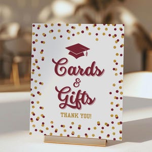 Könnte beinhalten: Ein Abschluss-Schild mit burgunderfarbenem Text "Cards & Gifts" und einem Mortarboard-Symbol. Das weiße Schild hat goldene und burgunderfarbene Konfetti, mit "THANK YOU!" unten. Es wird von einem goldenen Ständer gehalten.