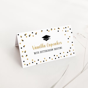 May include: A white folded tent card featuring a black graduation cap icon, gold script text reading Vanilla Cupcakes, and black text reading WITH BUTTERCREAM FROSTING. The card is decorated with a border of scattered black and gold confetti dots.