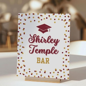 Puede incluir: Un letrero blanco con las palabras "Shirley Temple BAR" en letras burdeos y doradas, coronado con un gráfico de birrete burdeos. Puntos de confeti dorados y burdeos bordean el marco. El letrero está sujeto por un soporte dorado.