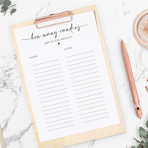 May include: A white paper game sheet on a wooden clipboard with the text How many candies ARE IN THE BOTTLE? and two columns for Name and Guess. The sheet features black text and lines, accompanied by a rose gold pen and small metal clips.