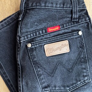 May include: Dark gray Wrangler jeans with a classic five-pocket design. Features a red Wrangler label and a tan leather patch with the brand name. The jeans are made of durable denim.