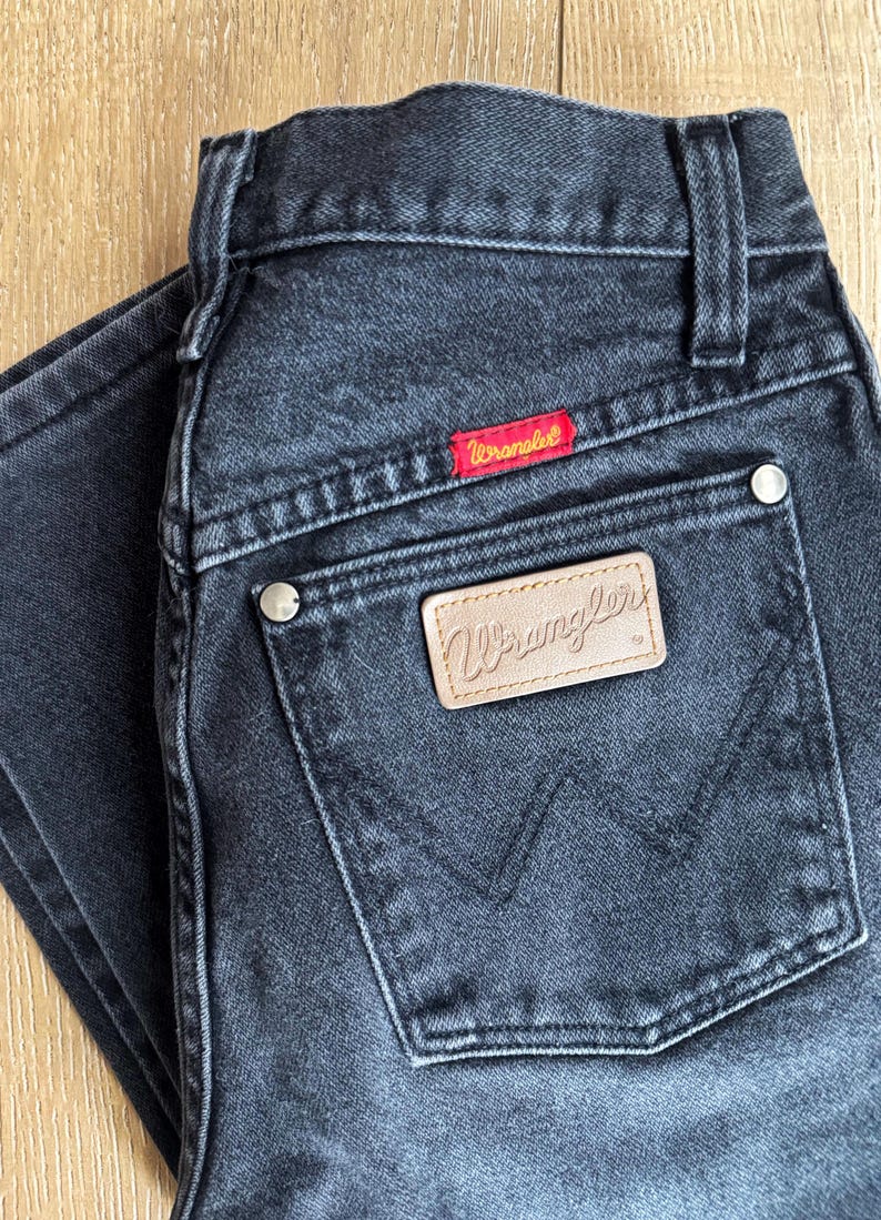 May include: Dark gray Wrangler jeans with a classic five-pocket design. Features a red Wrangler label and a tan leather patch with the brand name. The jeans are made of durable denim.