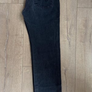 May include: Dark wash denim jeans with a classic five-pocket design. Features a brown leather patch and a red logo tag. The jeans are laid flat on a wooden surface.