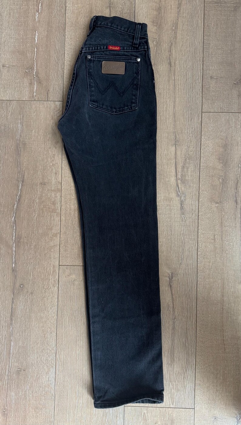 May include: Dark wash denim jeans with a classic five-pocket design. Features a brown leather patch and a red logo tag. The jeans are laid flat on a wooden surface.