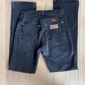 May include: A pair of black Wrangler jeans, laid flat. The jeans feature classic five-pocket styling, a leather patch with the Wrangler logo, and a red tag. The denim appears to be pre-worn, with subtle fading. The jeans are displayed on a light wood surface.