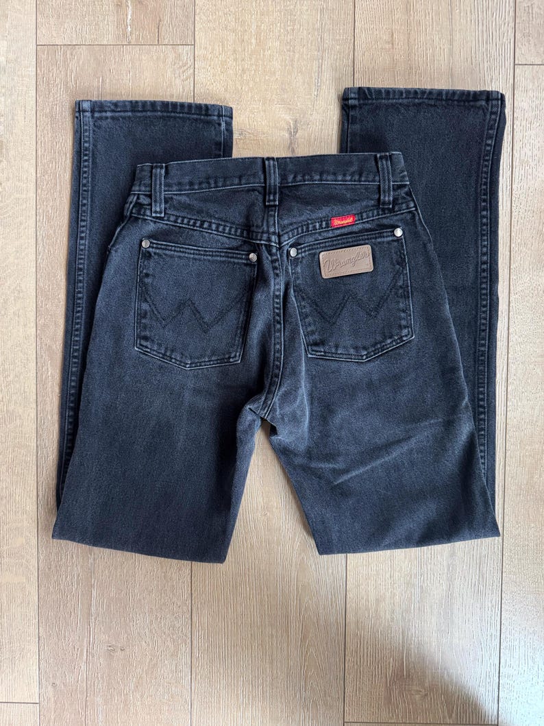 May include: A pair of black Wrangler jeans, laid flat. The jeans feature classic five-pocket styling, a leather patch with the Wrangler logo, and a red tag. The denim appears to be pre-worn, with subtle fading. The jeans are displayed on a light wood surface.