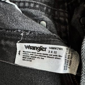May include: Close-up of a pair of black Wrangler jeans. The label reads "Wrangler 14MWZWK 3 X 32" and includes washing instructions. The jeans are made of 100% cotton and made in Mexico.