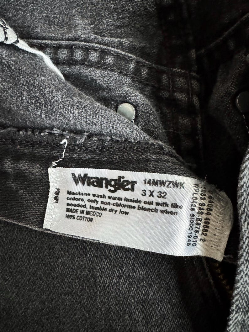 May include: Close-up of a pair of black Wrangler jeans. The label reads "Wrangler 14MWZWK 3 X 32" and includes washing instructions. The jeans are made of 100% cotton and made in Mexico.