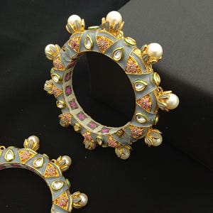 Peut inclure: Deux bracelets gris avec des détails dorés, ornés de motifs floraux roses, de perles blanches et de pierres précieuses transparentes en forme de goutte d'eau. Les bracelets présentent un design complexe et sont posés sur un fond noir.