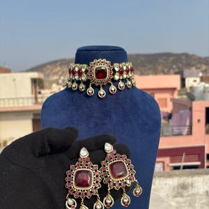 May include: A gold-toned choker necklace and matching earrings set. The jewelry features large, square, ruby-red center stones surrounded by smaller red and white stones. The earrings and necklace have dangling teardrop-shaped accents. The set is displayed on a blue mannequin head.