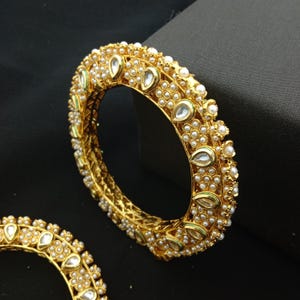 May include: Gold-toned bangle bracelet with a detailed design. The bracelet is adorned with small, round pearls and teardrop-shaped clear stones. The edges feature a delicate pattern, creating an elegant and ornate look. The jewelry is set against a dark background.
