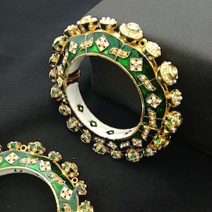 Puede incluir: Brazaletes dorados ornamentados con esmalte verde y detalles blancos. Los brazaletes presentan diseños intrincados con pequeñas decoraciones similares a perlas. Las joyas se presentan sobre un fondo oscuro.