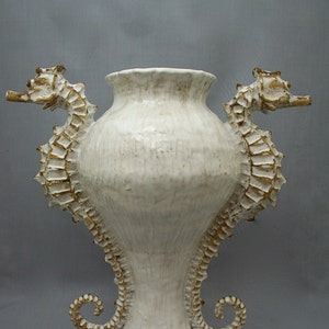 May include: White ceramic vase with two seahorse handles. The vase has a textured surface and is decorated with gold accents.