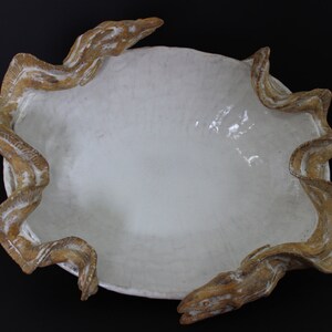 May include: A white ceramic bowl with a unique design. The bowl has a wavy, organic rim that resembles a series of flowing waves. The rim is a light brown color, giving the bowl a rustic look.