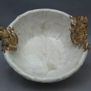 May include: White ceramic bowl with two frog-shaped handles. The bowl has a textured surface and a slightly off-white color.