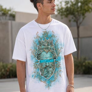 May include: White t-shirt featuring a detailed turquoise and gold graphic of a classical bust with a crown. The bust has a blue band across the eyes with the word "Neptune" printed on it. The shirt has short sleeves and a relaxed fit.