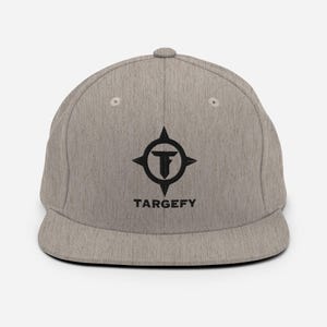 Puede incluir: Una gorra de béisbol gris jaspeado con visera plana y corona estructurada. La parte frontal presenta un logotipo bordado en negro con un diseño de brújula estilizada que rodea la letra T, con el texto TARGEFY en letras mayúsculas negras.