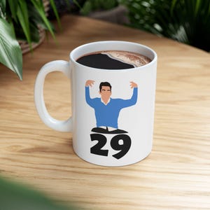 Può includere: Tazza da caffè in ceramica bianca con un'illustrazione a cartoni animati di una persona con un maglione blu con le braccia alzate. Sotto l'illustrazione c'è il numero 29 in nero. La tazza è piena di caffè scuro.