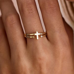 May include: A gold-colored ring with a cross design. The ring is worn on a finger, and the cross is the focal point. The ring has a delicate, elegant appearance. A small, clear stone is set next to the cross.