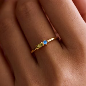 May include: A gold ring with a thin band and three gemstones. The ring has a small, rectangular light green stone, a small, round blue stone, and a small, rectangular light blue stone. The ring is on a person's finger.
