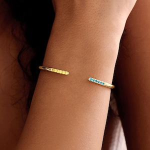 May include: A gold-coloured open cuff bracelet. One end is adorned with small yellow stones, and the other with small blue stones. The bracelet is worn on a person's wrist.