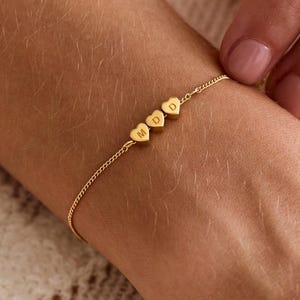 May include: A delicate gold bracelet featuring three heart-shaped charms, each engraved with a letter. The bracelet is worn on a wrist. The background is a soft, textured white fabric.