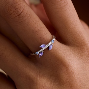 May include: A silver ring with a leaf motif, set with small, oval-cut purple gemstones and a few smaller blue stones. The ring is displayed on a finger. The design is elegant and nature-inspired.
