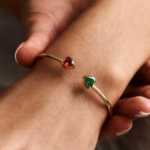 May include: Gold-coloured open cuff bracelet featuring heart-shaped gemstones. A red and a green gemstone are set at the ends of the bracelet. A gold ring with two heart-shaped gemstones is also visible.