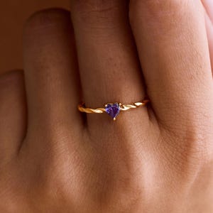 May include: A gold ring featuring a heart-shaped purple gemstone. The ring has a twisted band design. The ring is on a person's finger.