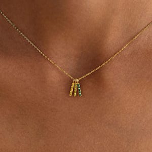 May include: A gold chain necklace featuring three small, vertical bar pendants. Each bar is encrusted with tiny, sparkling gemstones in distinct colours: one yellow, one orange, and one green, hanging together from the centre of the delicate chain.