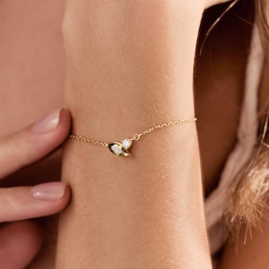 May include: A delicate gold bracelet with a small charm featuring a white gemstone and a green gemstone. A matching gold necklace is also visible, along with a gold ring. The jewellery is displayed on a person's arm.