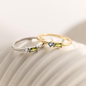 May include: Two thin rings, one silver-toned and one gold-toned, each featuring a small round gemstone and a rectangular baguette-cut gemstone. The silver ring has a clear stone and a green stone, while the gold ring has a blue and a green stone.