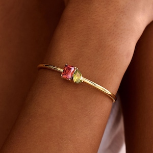 May include: A gold bracelet with a pink rectangular gemstone and a green pear-shaped gemstone. The bracelet is displayed on a wrist, highlighting the jewellery's design and colour contrast. The gemstones are the focal point.