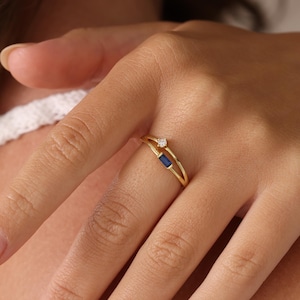 May include: A gold ring featuring a delicate double band design. One band is set with a small, round, clear gemstone, while the other band features a rectangular, deep blue sapphire-coloured stone. The metal has a polished, reflective finish.