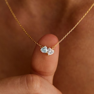Könnte beinhalten: Eine goldene Halskette mit zwei kleinen herzförmigen Anhängern. Ein Anhänger ist ein hellblauer Stein, der andere ein klarer, funkelnder Stein. Beide Herzen sind in goldenes Metall gefasst und an einer zarten, dünnen Goldkette befestigt.