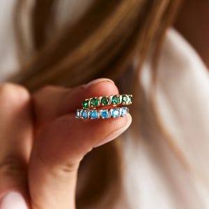 May include: Two gold-toned eternity rings, one with emerald green stones and the other with light blue stones. The rings are held between fingers, showcasing the jewelry's design and color contrast. The rings are a delicate accessory.