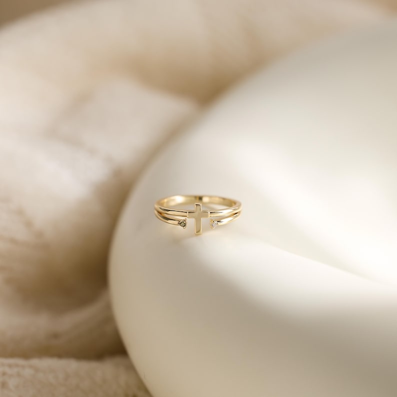May include: A gold ring featuring a central cross design flanked by two small, clear stones. The band is composed of two thin, parallel gold wires that meet at the cross, creating a delicate and open structure with a polished metallic finish.