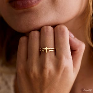 May include: A gold-toned open ring featuring a central cross design. The band is split at the front, with each end terminating in a small, clear, round-cut stone. The metal has a polished finish, and the cross is positioned vertically.